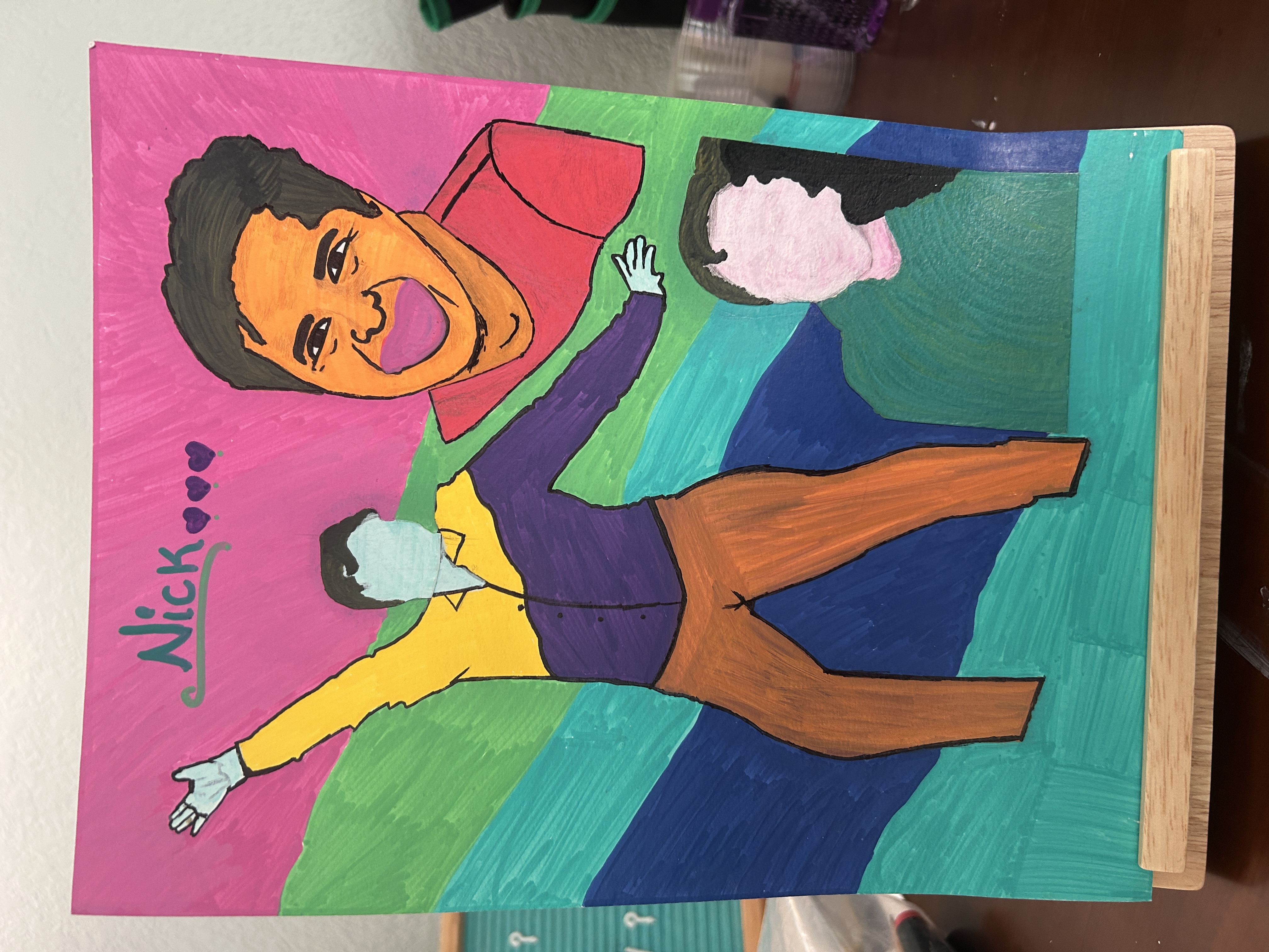 Colorful handmade portrait artwork displayed upright on a small wooden stand. The piece features bold pink, teal, green, and blue wavy background stripes with three stylized human figures outlined in black. One large smiling portrait appears in the upper right, while two faceless figures appear in the center and lower right. The name “Nick” with three hearts is written across the top in green and purple lettering. The artwork has a vibrant, expressive, deeply personal feel with visible marker and brush strokes throughout.