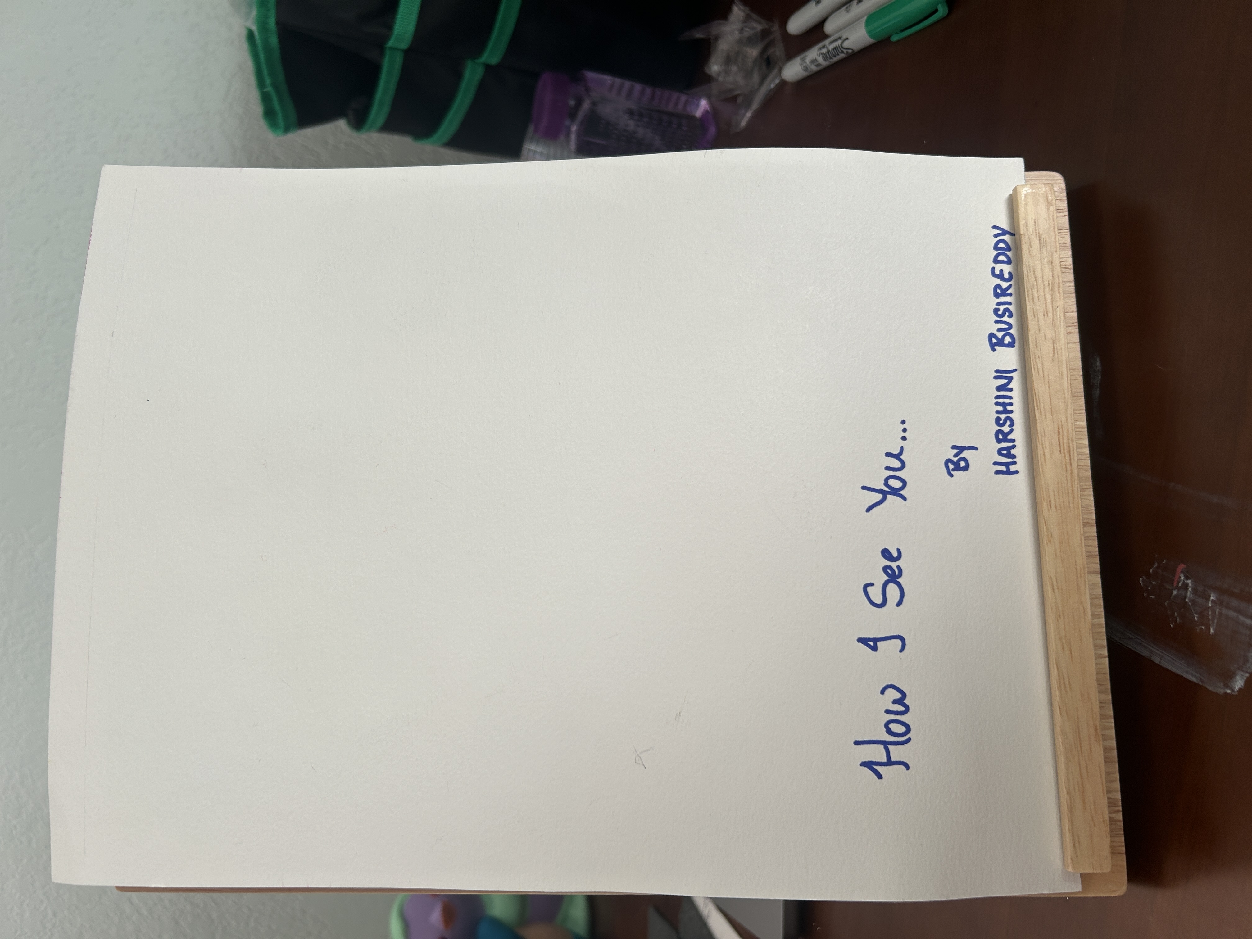Back view of a handmade portrait artwork displayed upright on a small wooden stand. The back of the paper is mostly blank white with handwritten blue text near the bottom reading, “How I See You…” and underneath, “By Harshini Busireddy.” The artwork sits against a softly lit indoor background with art supplies partially visible nearby.