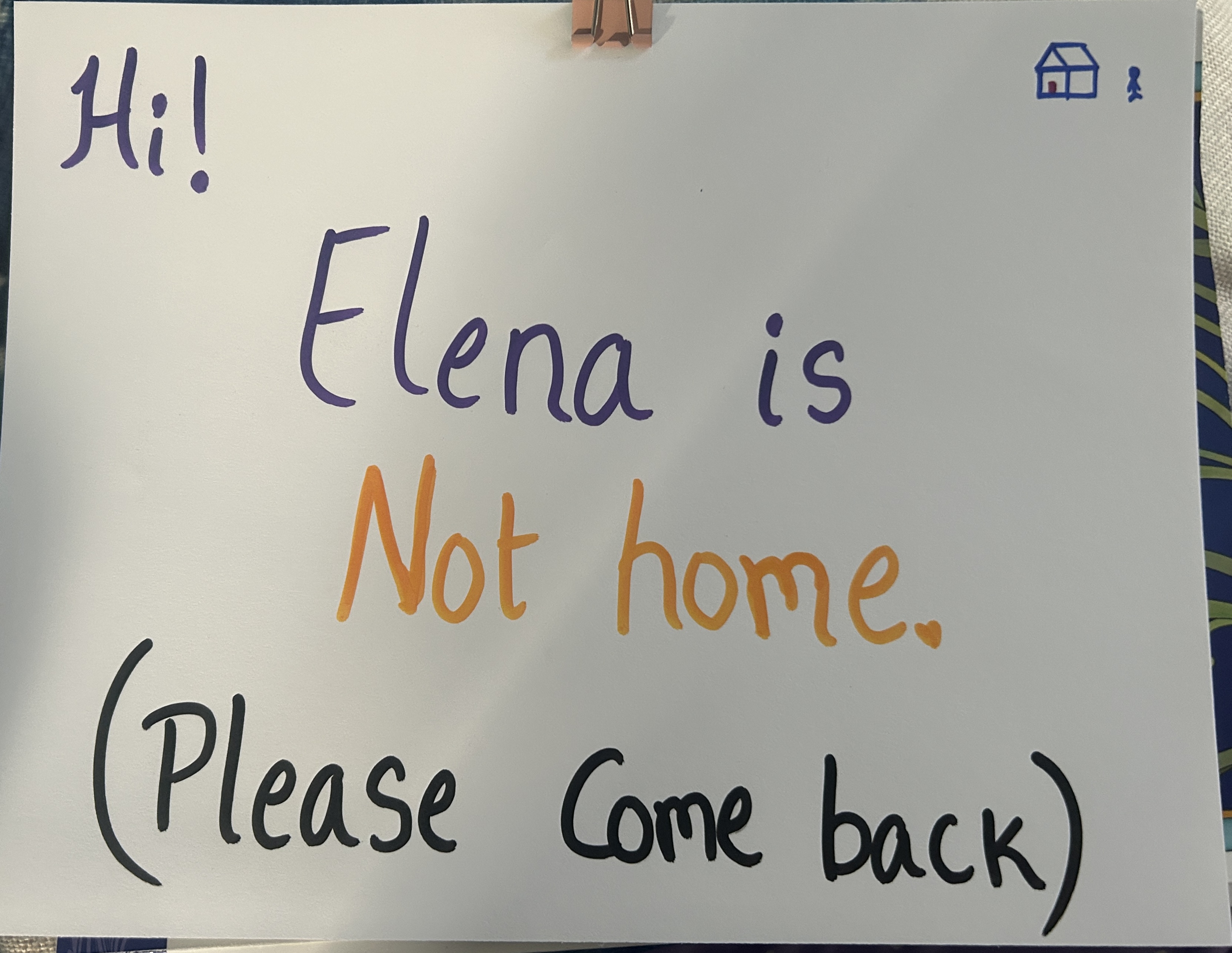 A handwritten door sign clipped to a wall reading "Hi! Elena is Not home. (Please Come back)" in purple, orange, and black marker, with small house and figure icons in the corner.