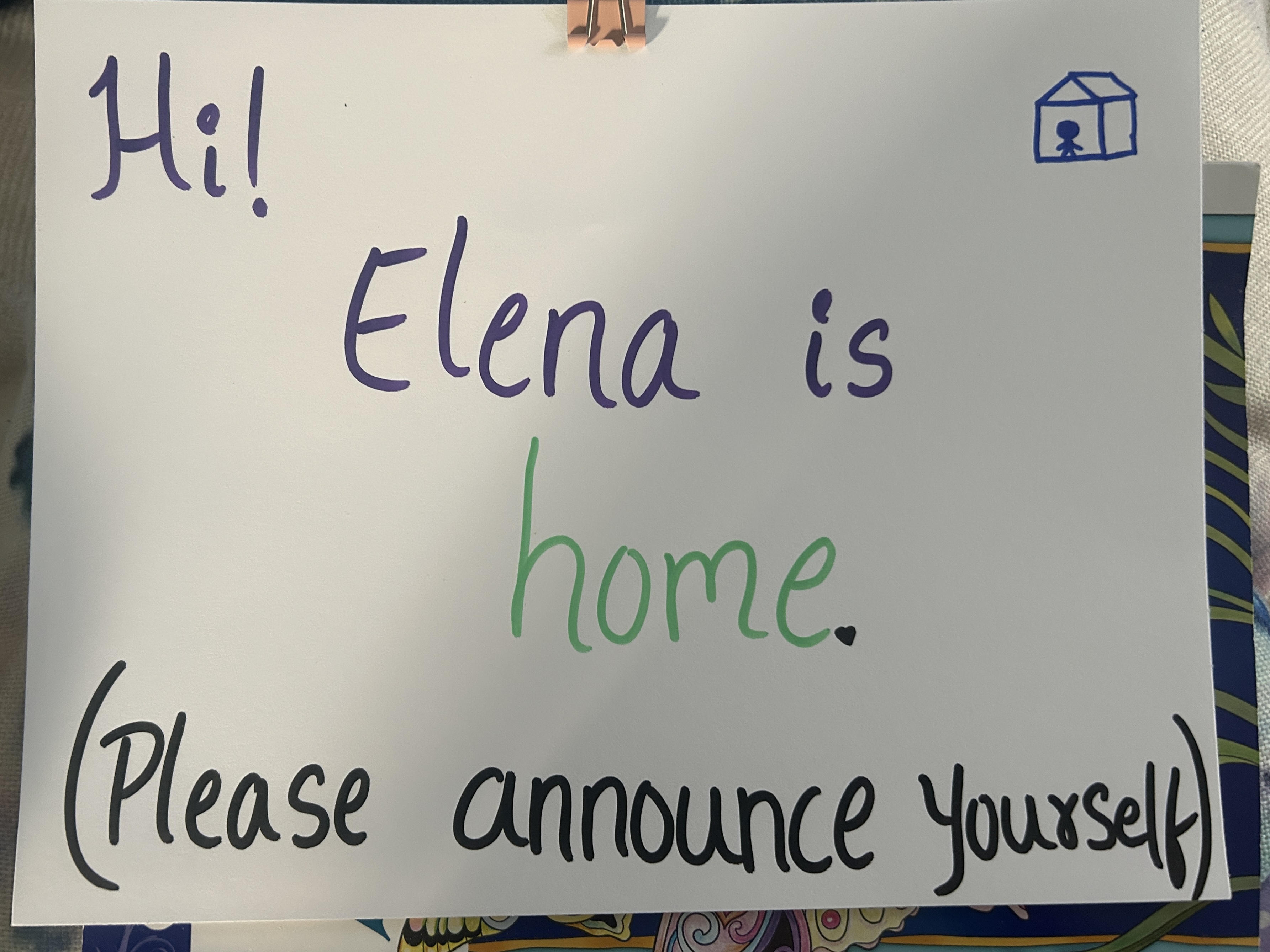 A handwritten door sign clipped to a wall reading "Hi! Elena is home. (Please announce yourself)" in purple, green, and black marker, with a small house icon showing a figure inside.
