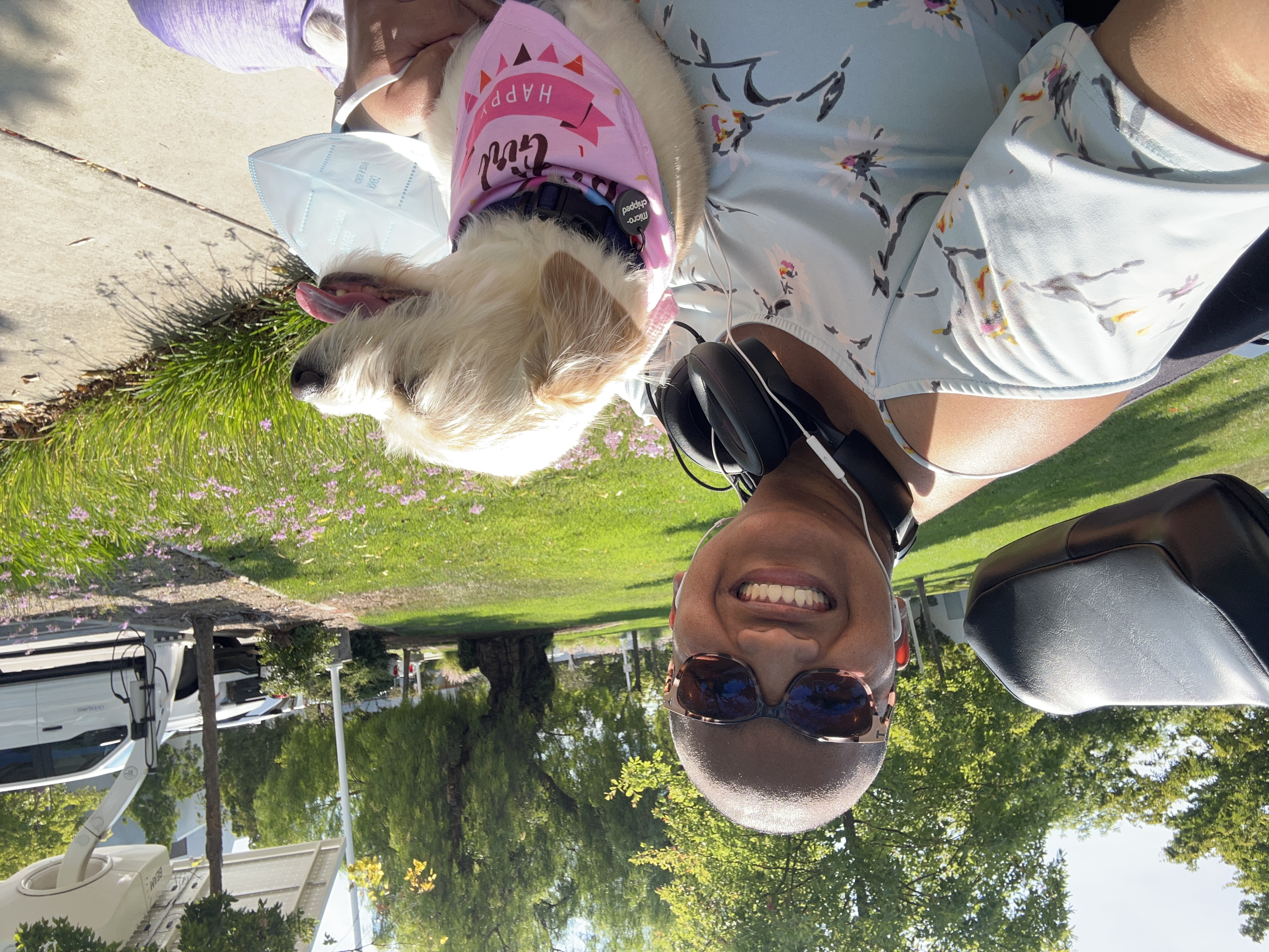 Elena smiles beside Toffee outdoors on a sunny day. Toffee is seated with her on a power wheelchair and wearing a pink birthday bandana.