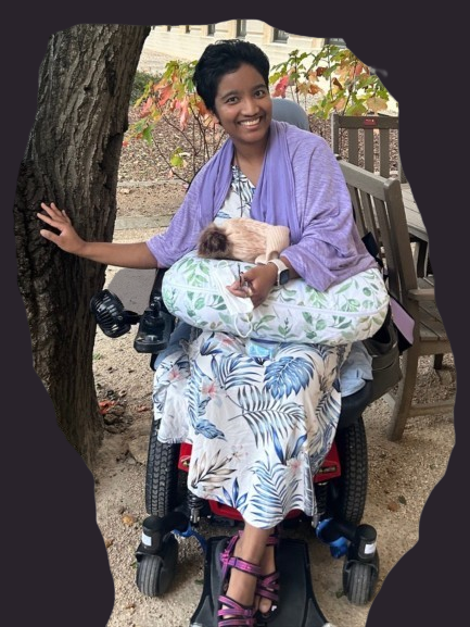 Harshini Busireddy (now Elena Claire) smiling outdoors in a power wheelchair with a small animal resting on a pillow in her lap.