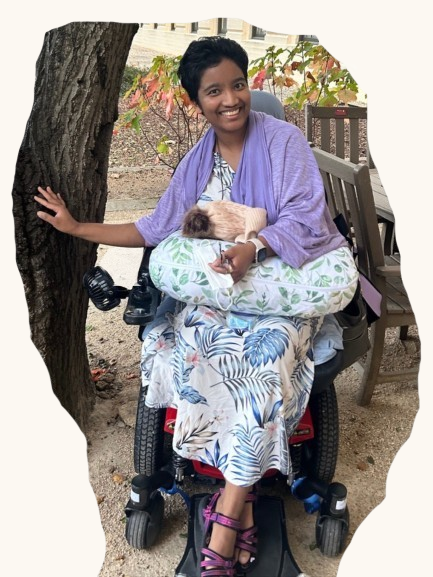 Harshini Busireddy (now Elena Claire) smiling outdoors in a power wheelchair with a small animal resting on a pillow in her lap.