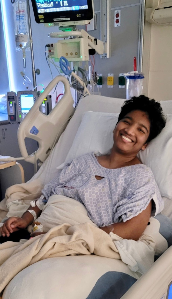 Harshini smiling in a hospital bed wearing a hospital gown with medical equipment in the background.