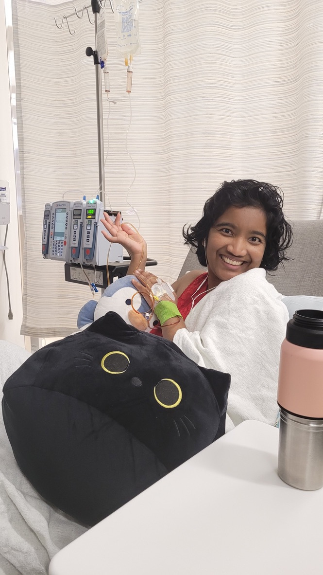 Harshini smiling while seated in hospital chair with IV lines, holding a black cat plush.