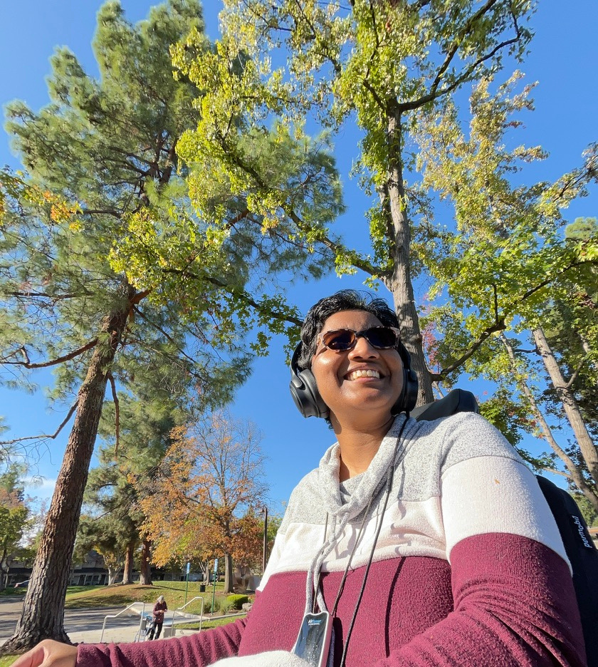 Harshini outdoors under tall trees, smiling and looking upward.